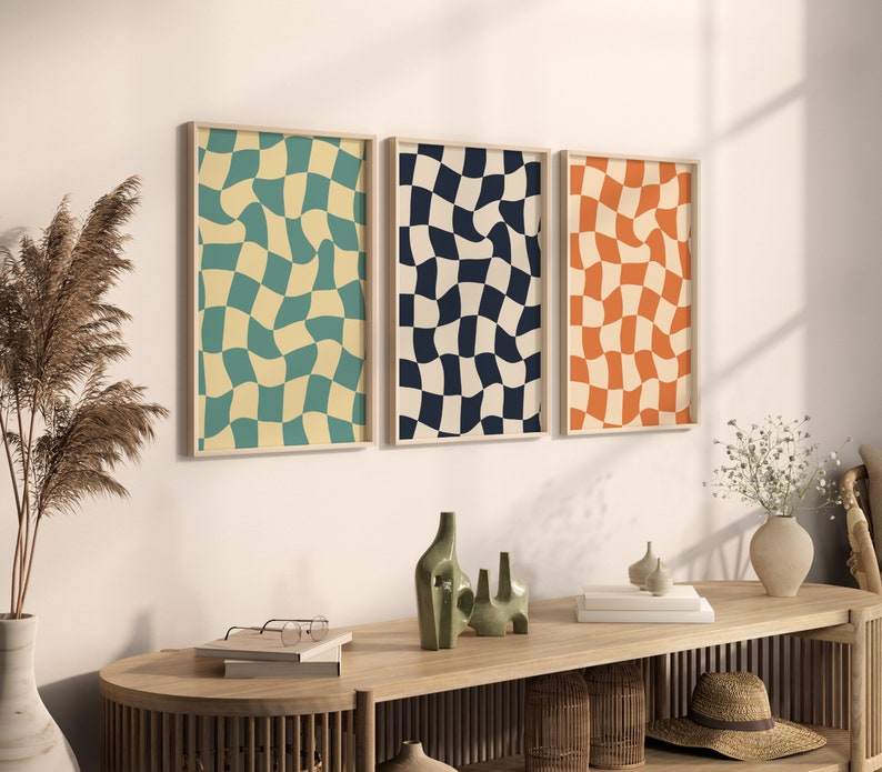 May include: Three framed abstract prints with a checkered pattern. The prints are in shades of blue, green, and orange. The prints are hanging on a white wall above a wooden table.