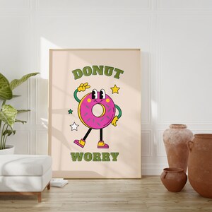 May include: A pink donut with green sprinkles and a smiling face is depicted on a beige poster with the text "DONUT WORRY" in green letters. The poster is framed in a light brown frame and is hanging on a white wall.