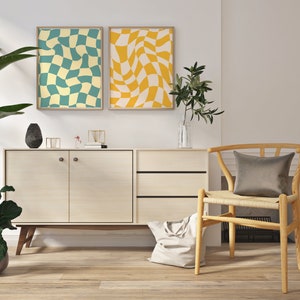 May include: Two abstract geometric prints in yellow and blue hang above a light wood cabinet with two doors and two drawers. A woven chair with a gray cushion sits to the right of the cabinet.