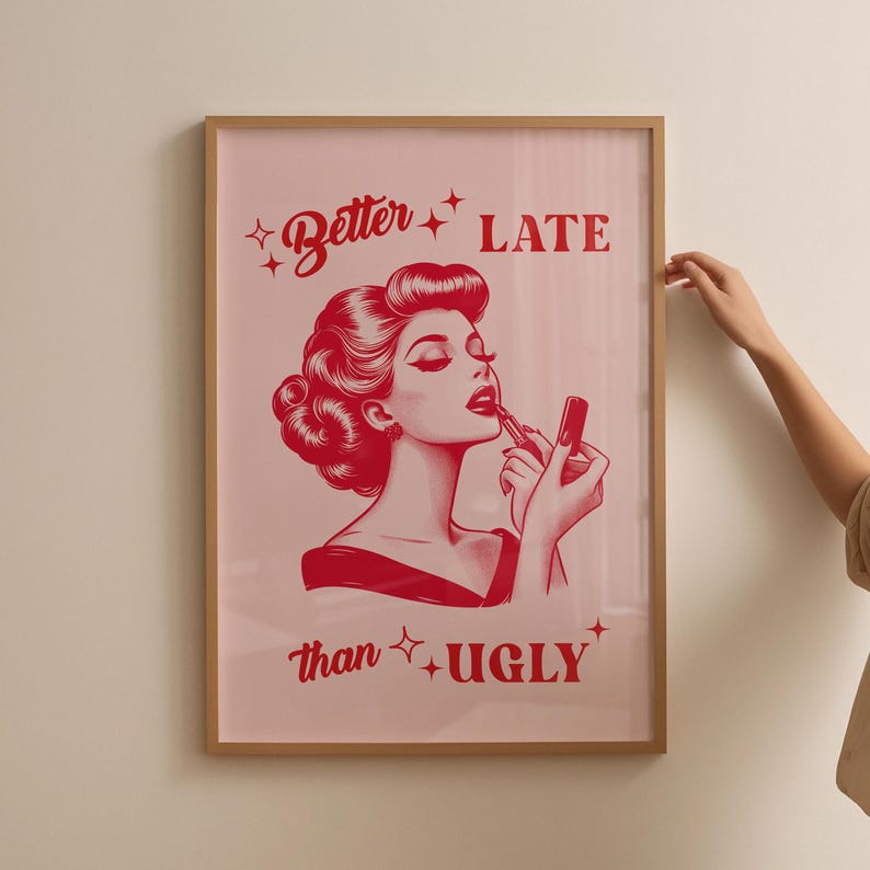 Pu&ograve; includere: Una stampa incorniciata con sfondo rosa e testo rosso che recita "Better Late than Ugly". Al centro della stampa &egrave; presente un'illustrazione rossa di una donna che si applica il rossetto.