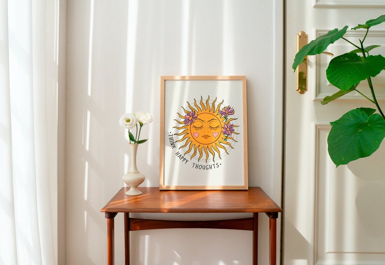 Puede incluir: Una impresi&oacute;n enmarcada con un sol amarillo con una cara y los ojos cerrados. El sol est&aacute; rodeado de flores rosas y el texto "Think Happy Thoughts." est&aacute; escrito debajo del sol.