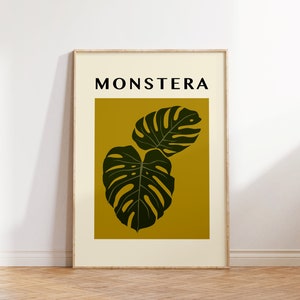 May include: A framed print with the word "MONSTERA" in black text on a white background. The print features two dark green monstera leaves on a yellow background.