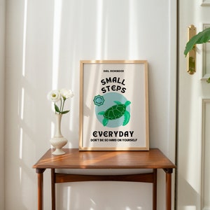 Pu&ograve; includere: Una stampa incorniciata con una cornice marrone chiaro e uno sfondo bianco. La stampa presenta una tartaruga marina verde con il testo "Small Steps Everyday Don't be so hard on yourself."