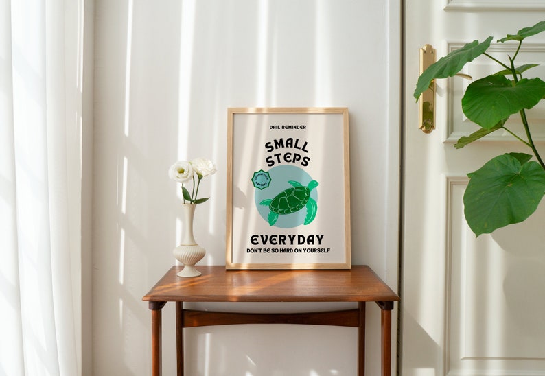 Pu&ograve; includere: Una stampa incorniciata con una cornice marrone chiaro e uno sfondo bianco. La stampa presenta una tartaruga marina verde con il testo "Small Steps Everyday Don't be so hard on yourself."