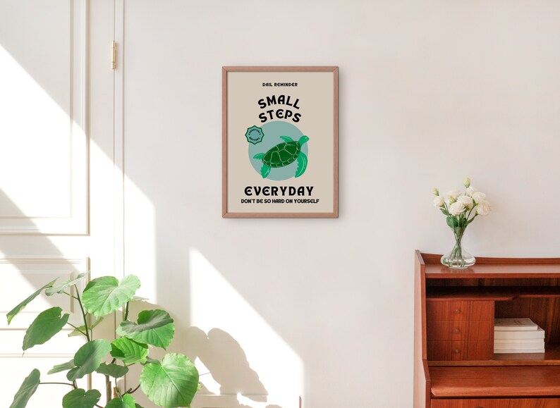 Pu&ograve; includere: Un poster motivazionale incorniciato con sfondo blu chiaro e una tartaruga marina verde. Il poster recita "Small Steps Everyday Don't be so hard on yourself".