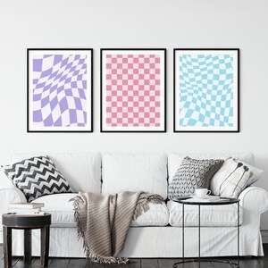 May include: Three framed prints of checkered patterns in a living room setting. The prints are in shades of purple, pink, and blue. The prints are hanging on a white wall above a white couch with pillows and a throw blanket.