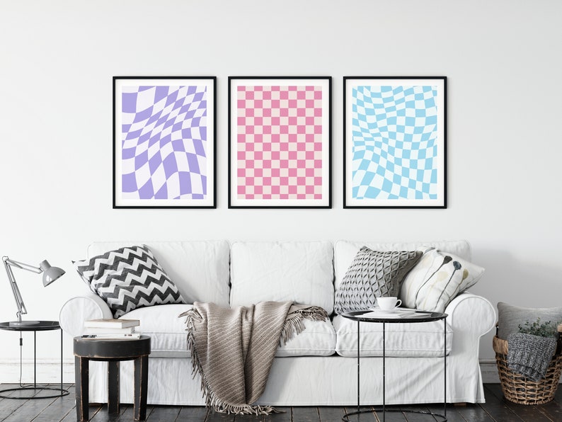 May include: Three framed prints of checkered patterns in a living room setting. The prints are in shades of purple, pink, and blue. The prints are hanging on a white wall above a white couch with pillows and a throw blanket.