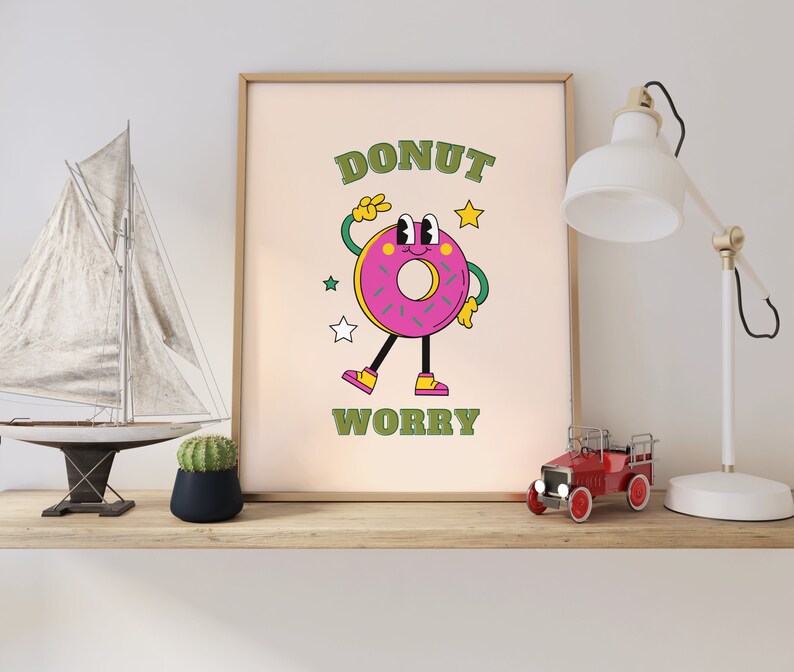 May include: A framed print with a cartoon donut character with the text "DONUT WORRY" in green letters. The donut is pink with green sprinkles and a smiling face. The background is a light beige color.