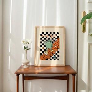 May include: A framed print with a black and white chequered background. A green frog is walking up a set of orange stairs. The text "Frog You!" is written in black letters on the print.