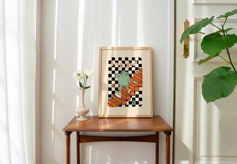May include: A framed print with a black and white chequered background. A green frog is walking up a set of orange stairs. The text "Frog You!" is written in black letters on the print.