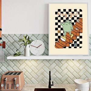 May include: A white framed print featuring a green frog wearing shoes, ascending a staircase with a checkered pattern. The text "Frog you!" is written in orange and brown letters.