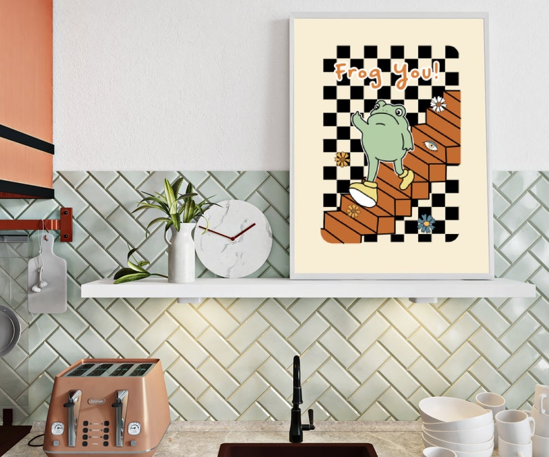 May include: A white framed print featuring a green frog wearing shoes, ascending a staircase with a checkered pattern. The text "Frog you!" is written in orange and brown letters.