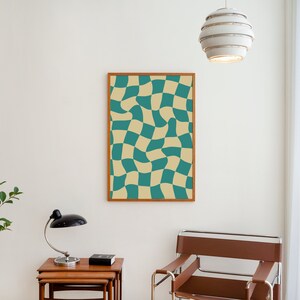 May include: A framed print of a checkered pattern with teal and beige squares. The squares are distorted, creating a wavy effect. The print is hanging on a white wall in a room with a wooden table, a lamp, and a chair.