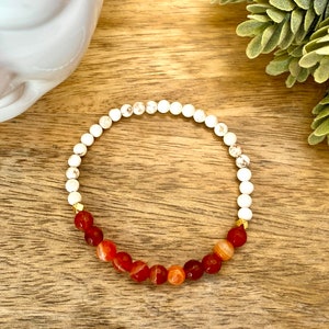 May include: A white and red beaded bracelet with gold accents. The bracelet is made with white howlite beads and red carnelian beads.