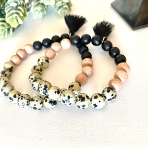 May include: Two beaded bracelets. One bracelet features Dalmatian jasper beads, light wood beads, and black beads. The other bracelet has the same design. Each bracelet has a black tassel.