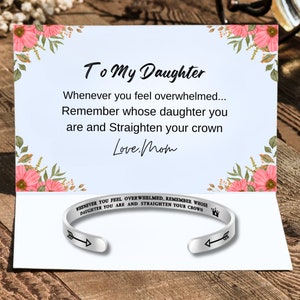 Può includere: Un braccialetto d'argento con un design a freccia e corona. Il braccialetto ha un'iscrizione che recita "Whenever you feel overwhelmed, remember whose daughter you are and straighten your crown". Il braccialetto è accompagnato da una carta con un design floreale e il messaggio "To My Daughter, Whenever you feel overwhelmed... Remember whose daughter you are and straighten your crown. Love, Mom."