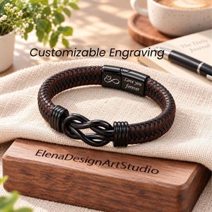 May include: A braided dark brown leather bracelet featuring a black metal infinity knot centrepiece and a black clasp engraved with an infinity symbol, a heart, and the text Love you forever. The bracelet rests on a textured fabric surface.
