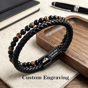 May include: A multi-strand bracelet featuring a braided black leather cord and a string of polished black and brown tiger eye beads. The bracelet includes a black metal magnetic clasp engraved with a mountain icon and the text Love you forever.