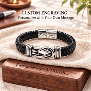 May include: A black braided leather bracelet featuring a silver-toned metal knot centerpiece and a clasp engraved with an infinity symbol, a heart, and the text Love you forever xx Nana. The bracelet is displayed on a textured fabric.