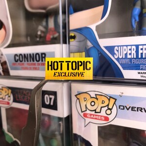 May include: A yellow sticker with the text "HOT TOPIC EXCLUSIVE" on a shelf with various Funko Pop figures. The figures include Connor from Assassin's Creed, Batman, and Overwatch characters.