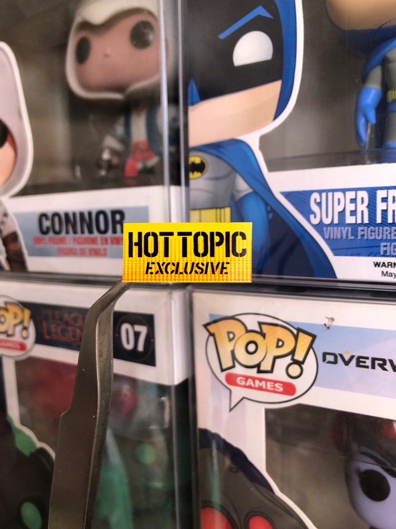 May include: A yellow sticker with the text "HOT TOPIC EXCLUSIVE" on a shelf with various Funko Pop figures. The figures include Connor from Assassin's Creed, Batman, and Overwatch characters.