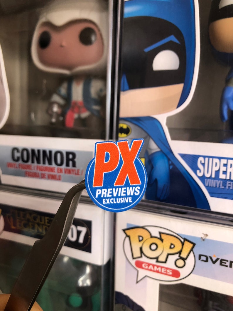 May include: A red and white circular sticker with the text "PX Previews Exclusive" on a white background. The sticker is attached to a clear plastic box with a Funko Pop! figure inside.