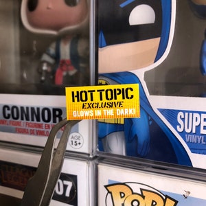 May include: Yellow sticker with the text "HOT TOPIC EXCLUSIVE GLOWS IN THE DARK!" on a black and blue background. The sticker is attached to a plastic container with a figure inside.