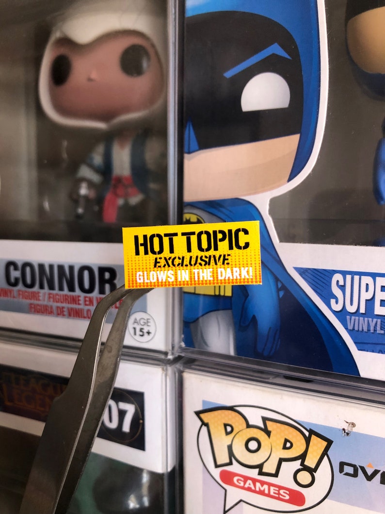 May include: Yellow sticker with the text "HOT TOPIC EXCLUSIVE GLOWS IN THE DARK!" on a black and blue background. The sticker is attached to a plastic container with a figure inside.