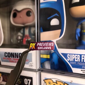 May include: A close-up of a red and yellow sticker that says "PX PREVIEWS EXCLUSIVE" on a pair of silver tweezers. The sticker is being held over a display of Funko Pop! figures.