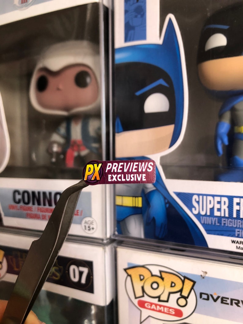 May include: A close-up of a red and yellow sticker that says "PX PREVIEWS EXCLUSIVE" on a pair of silver tweezers. The sticker is being held over a display of Funko Pop! figures.
