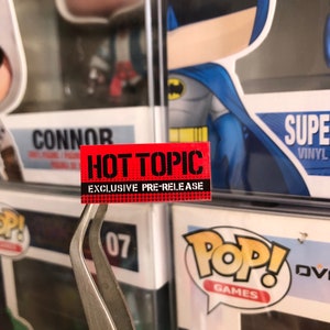 May include: A red and black sticker with the text "HOT TOPIC EXCLUSIVE PRE-RELEASE" on a white background. The sticker is being held by a pair of silver tweezers.