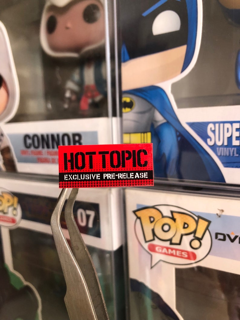 May include: A red and black sticker with the text "HOT TOPIC EXCLUSIVE PRE-RELEASE" on a white background. The sticker is being held by a pair of silver tweezers.