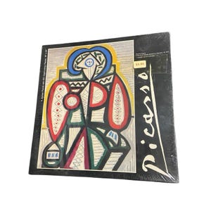 May include: A square jigsaw puzzle box featuring a Picasso painting titled Femme Assise. The box is black with white text reading Picasso and A Jigsaw Puzzle with 551 Interlocking Pieces, 18 x 24. The artwork displays abstract shapes in blue, red, green, and yellow.