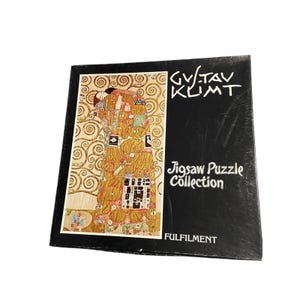 May include: A square jigsaw puzzle box featuring Gustav Klimt's The Kiss artwork on the left side. The right side is black with white text reading GVSTAV KLIMT, Jigsaw Puzzle Collection, and FULFILMENT printed at the bottom of the box.
