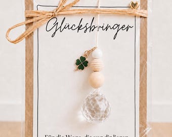 May include: A decorative hanging ornament featuring a clear faceted crystal bead, two wooden beads, and a small green four-leaf clover charm. It is attached to a white card with the text "Glücksbringer" and "Für die Wege, die vor dir liegen, und alles, was kommen mag" tied with a tan raffia bow.