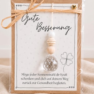 Könnte beinhalten: Eine Grußkarte in einer transparenten Kunststoffhülle mit Bastschleife, bedruckt mit dem Text Gute Besserung, einer vierblättrigen Kleeblatt-Zeichnung und einem Anhänger aus einer weißen Perle, zwei Holzperlen und einer facettierten Kristallkugel.