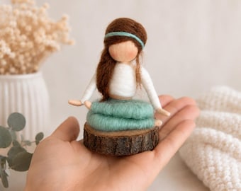 May include: A handmade felt doll with brown hair, a white top, and a teal skirt, seated on a wooden base. The doll has a teal headband and outstretched arms. The doll is held in a person's hand.