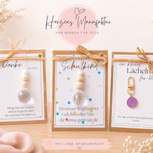 May include: Three gift cards with kraft paper borders, each tied with a raffia bow. Two cards feature a hanging crystal prism with wooden beads, while the third displays a purple smiley face charm. Each card includes German text and decorative elements.