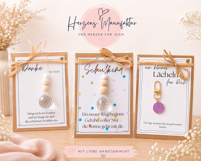 May include: Three gift cards with kraft paper borders, each tied with a raffia bow. Two cards feature a hanging crystal prism with wooden beads, while the third displays a purple smiley face charm. Each card includes German text and decorative elements.