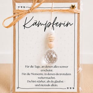 Könnte beinhalten: Eine rechteckige Karte mit Kraftpapier-Rahmen, die den deutschen Text KÄMPFERIN und ein motivierendes Zitat enthält. Ein hängendes Ornament mit einer klaren facettierten Kristallperle, zwei Holzperlen und einer weißen Perle ist befestigt. Eine Bastschleife und ein kleines goldenes Herz verzieren den oberen Teil.