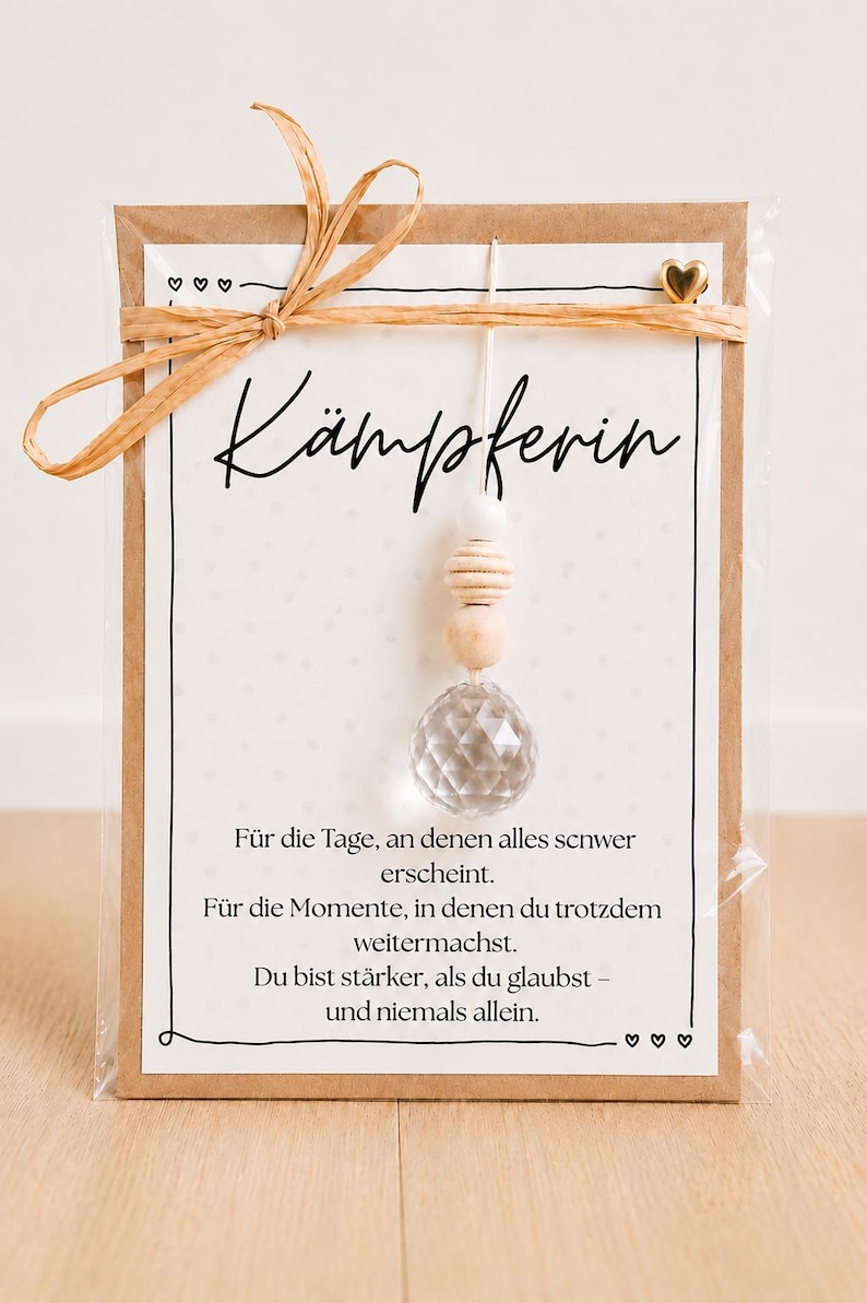 May include: A rectangular card with a kraft paper border featuring the German text K&Auml;MPFERIN and a motivational quote. A hanging ornament with a clear faceted crystal bead, two wooden beads, and a white bead is attached. A raffia bow and a small gold heart accent the top.