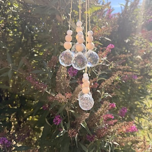May include: A hanging suncatcher ornament featuring three clear, multifaceted crystal glass spheres suspended by thin strings. Each sphere is topped with a vertical arrangement of smooth, round, light-coloured wooden beads of varying sizes.