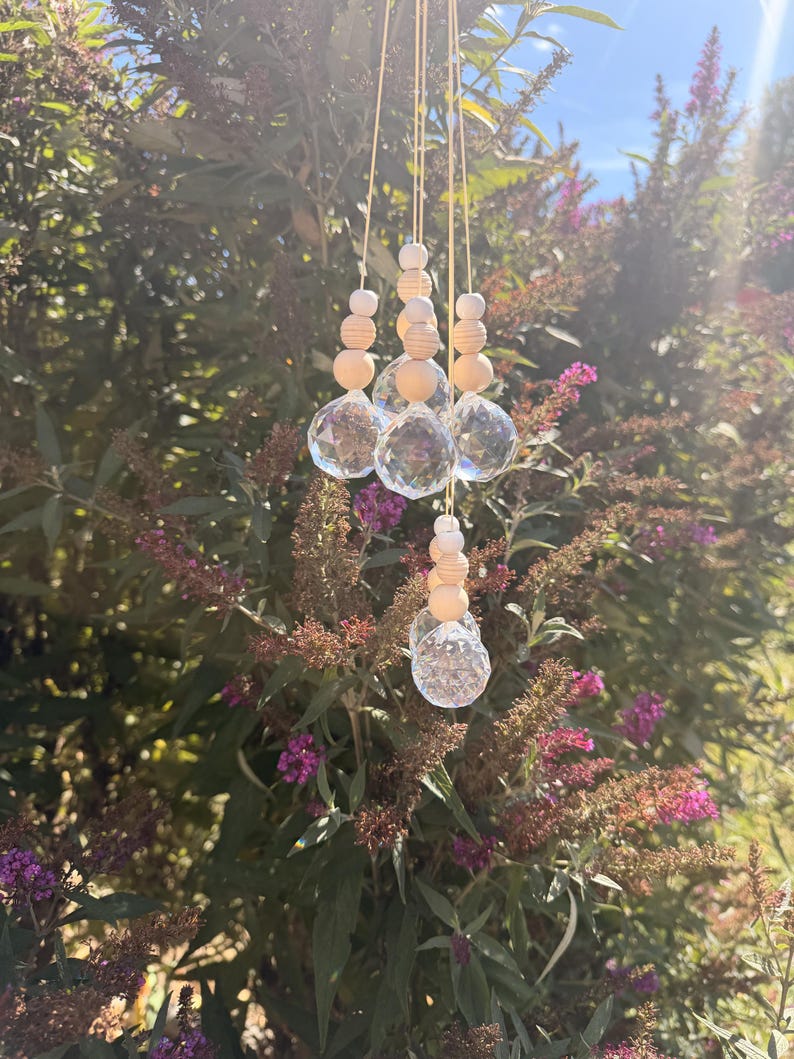 May include: A hanging suncatcher ornament featuring three clear, multifaceted crystal glass spheres suspended by thin strings. Each sphere is topped with a vertical arrangement of smooth, round, light-coloured wooden beads of varying sizes.