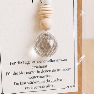 May include: A decorative hanging ornament featuring a faceted clear glass sphere suspended by a white cord with a white bead and two natural wooden beads. The ornament is attached to a card with the German text K&auml;mpferin and an inspirational message.