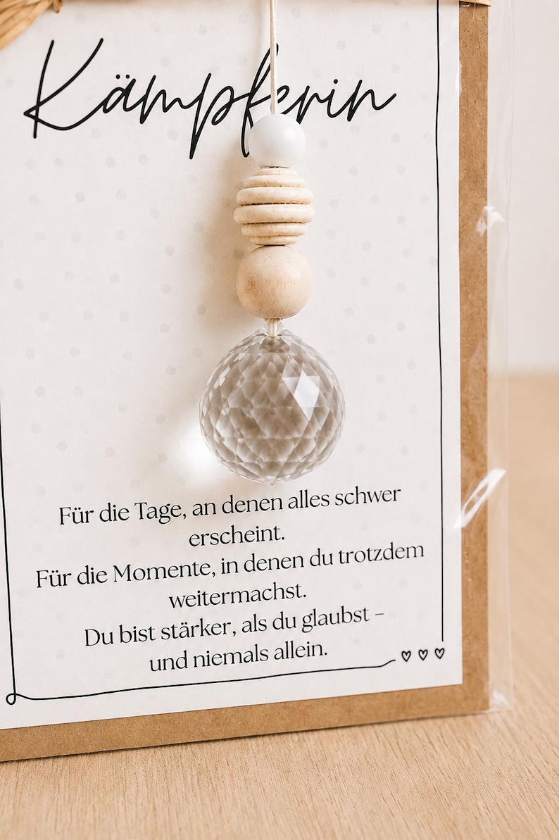 May include: A decorative hanging ornament featuring a faceted clear glass sphere suspended by a white cord with a white bead and two natural wooden beads. The ornament is attached to a card with the German text K&auml;mpferin and an inspirational message.