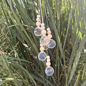 May include: A hanging ornament featuring five clear, multifaceted crystal spheres suspended by thin white cords. Each crystal is accented with a series of small, round, light-coloured wooden beads and smaller white beads arranged in a vertical pattern.