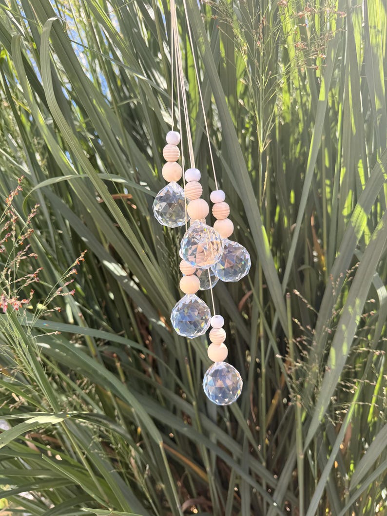 May include: A hanging ornament featuring five clear, multifaceted crystal spheres suspended by thin white cords. Each crystal is accented with a series of small, round, light-coloured wooden beads and smaller white beads arranged in a vertical pattern.