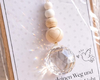 May include: A decorative hanging ornament featuring a clear faceted crystal sphere suspended by a string of wooden beads, including one with a heart engraving. The ornament is attached to a card with the text Zur Firmung and Gott segne deinen Weg und schenke dir immer sein Licht, decorated with a dove illustration and a small gold heart.