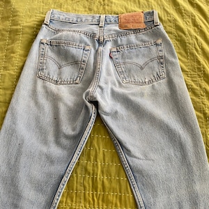May include: Light blue denim jeans with a faded wash and a brown leather patch with the text "Levi's" and "501" on the back. The jeans are laid flat on a green quilted fabric.