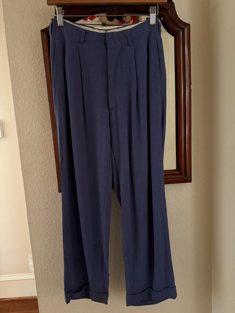 K&ouml;nnte beinhalten: Marineblaue Hose mit Falten, h&auml;ngt an einem Holzb&uuml;gel. Die Hose hat eine lockere Passform mit umgeschlagenem Saum. Die Hose ist aus leichtem Stoff und scheint in gutem Zustand zu sein.
